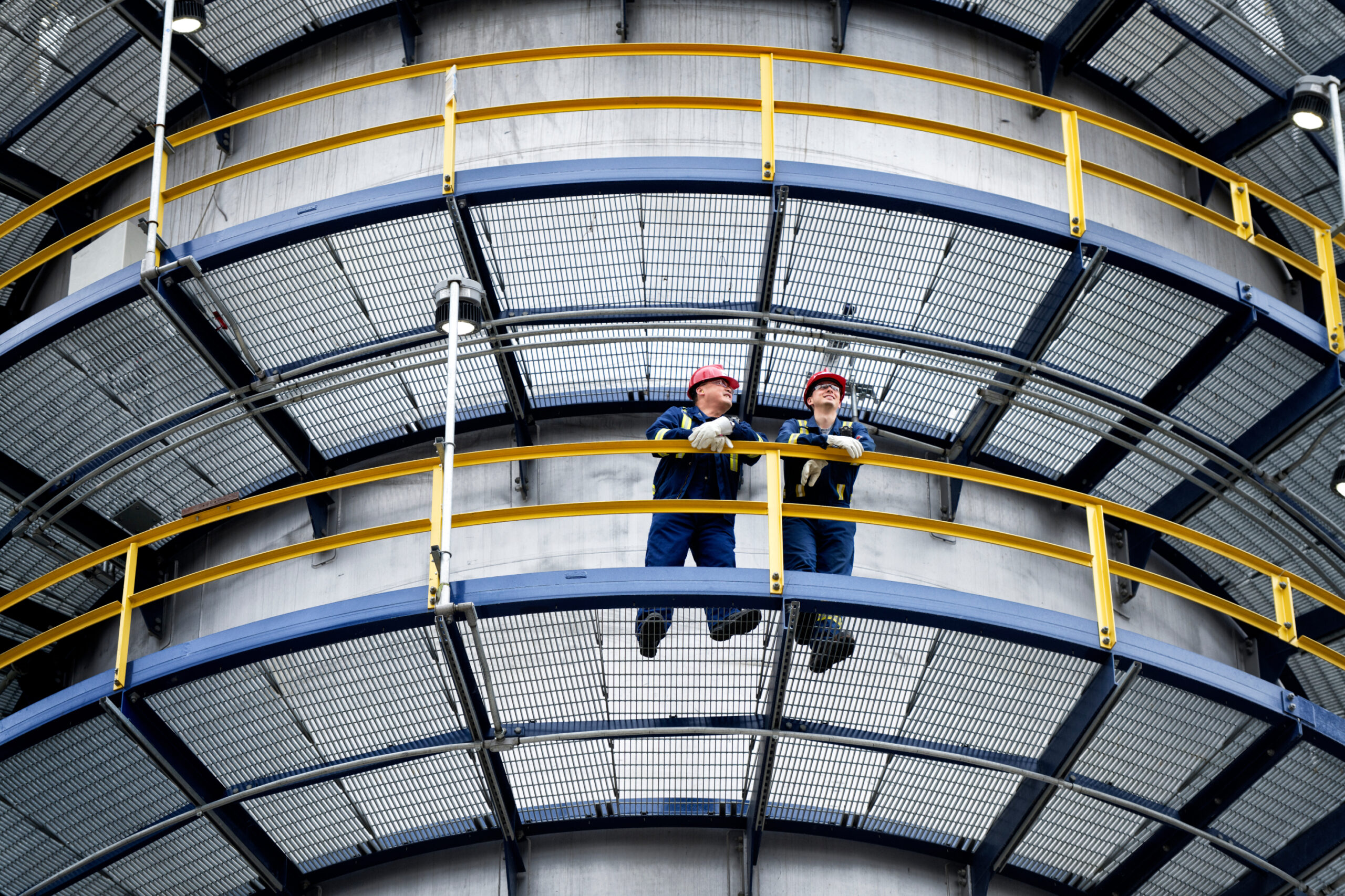 Two people talking at an operating facility. They're wearing red hard hats, gloves, protective safety glasses and blue uniforms with reflective stripes.