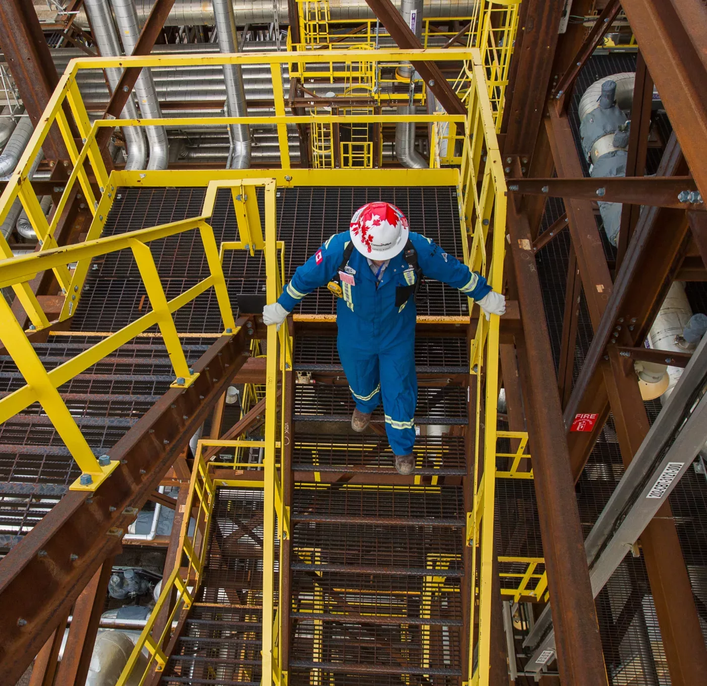 Person at an operating facility in protective gear, walking down metal stairs, viewed from above.
