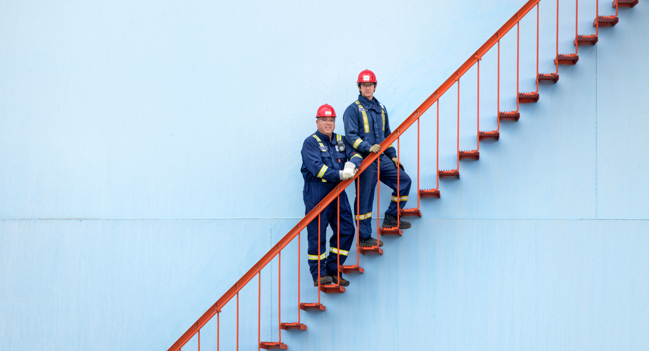 Two people wearing red hard hats and protective clothing standing on a red metal staircase at an operating facility.