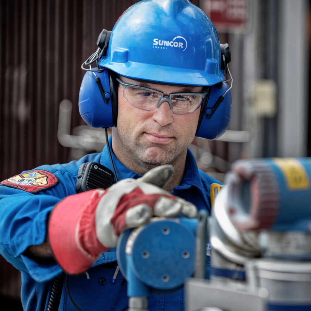 Close-up of person wearing protective glasses, blue hard hat and red gloves working at an operating facility.