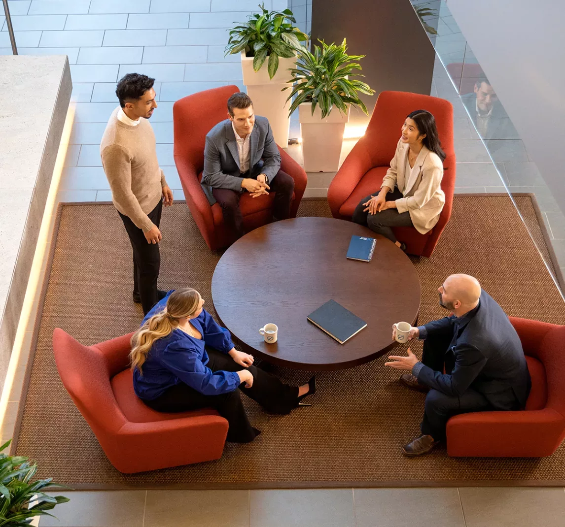 Cinq adultes dans un bureau. Ils sont réunis autour d’une petite table ronde avec des tasses à café. Quatre personnes sont assises autour de la table, et une personne est debout.