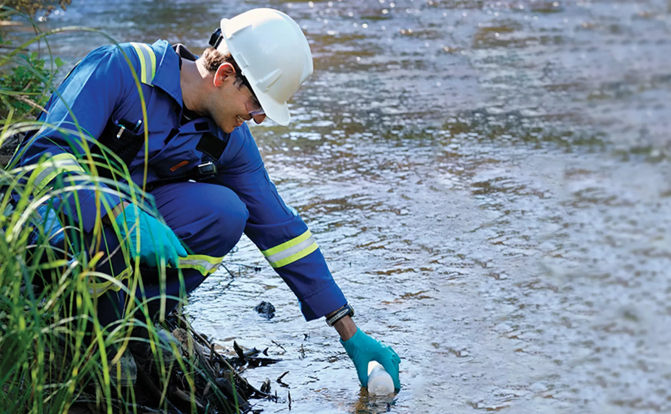 Person in blue uniform and gloves, crouching next to a river and retrieving a water sample.