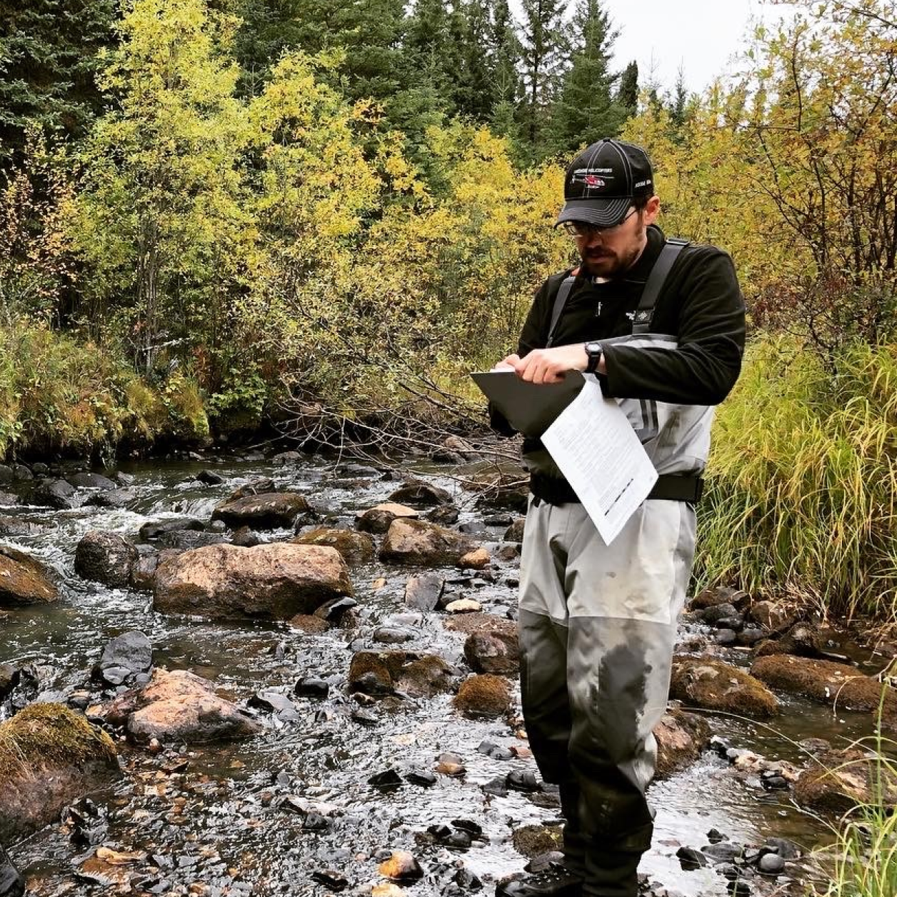 Person wearing glasses, a black cap, black shirt and light grey overalls standing in a shallow stream surrounded by rocks and trees. They are writing on a clipboard.