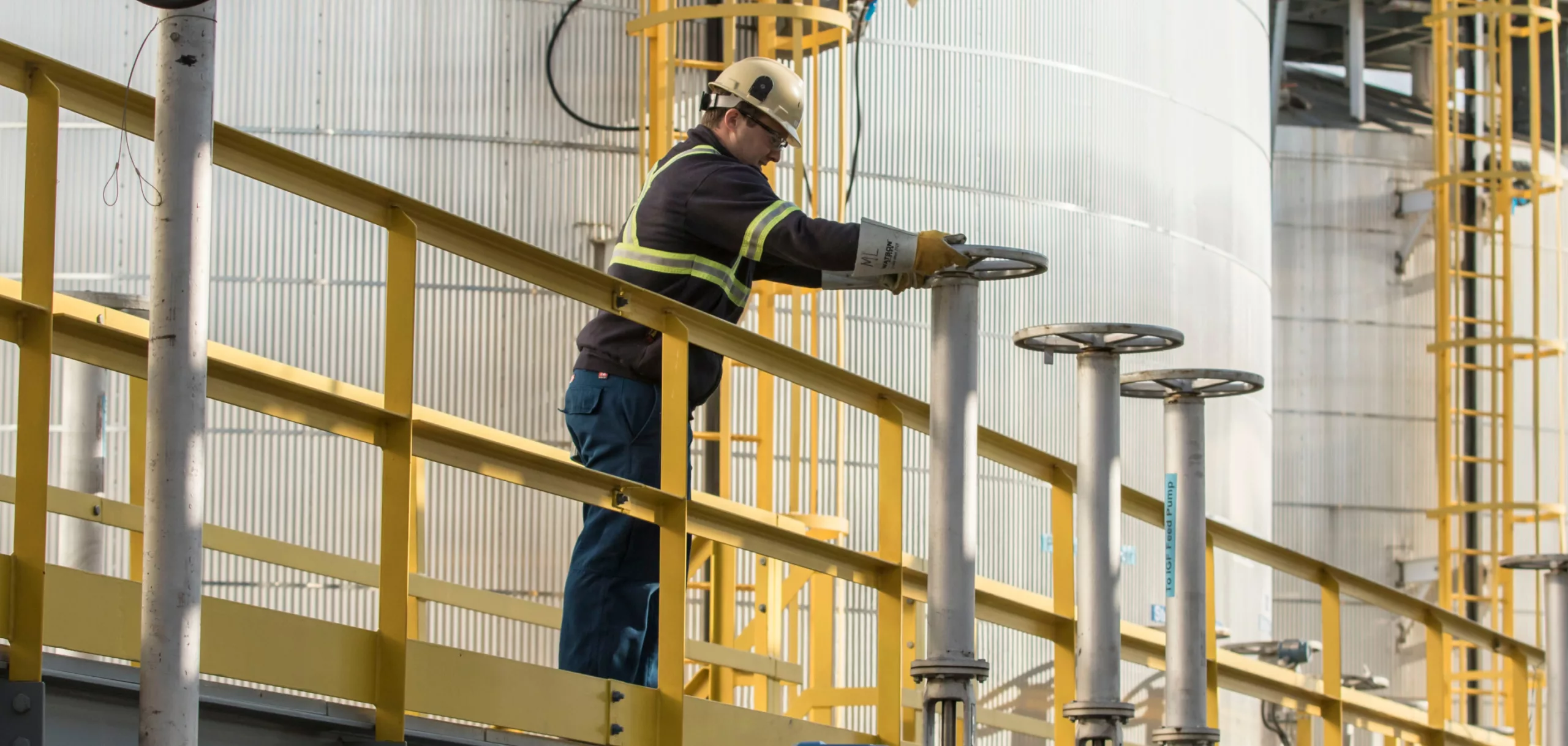 Person working outdoors at an operating facility, wearing a reflective black jacket, white hard hat and protective gloves.